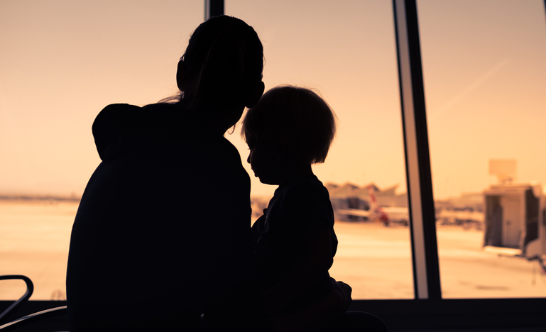 Silhouetted moment: a parent and child gaze out of a vast airport window, watching the bustle on the tarmac as they await their flight.