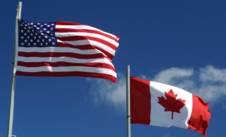Canadian and U.S. flag flying under a blue sky.