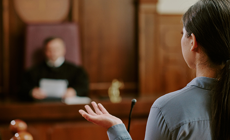 A woman speaks in a courtroom, facing a judge seated at a wooden bench. The scene conveys a serious and formal tone.