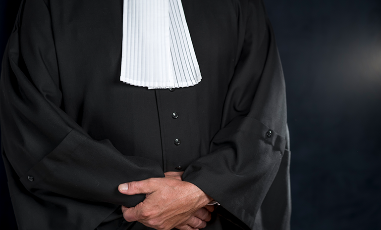 Person wearing a black legal robe and white court tabs, hands clasped in front.