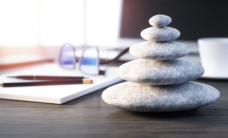 A stack of smooth gray stones sits on a desk, symbolizing balance. Nearby are glasses, a notebook, and a cup, creating a calm, organized workspace.