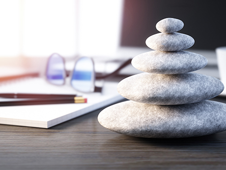 A stack of smooth gray stones sits on a desk, symbolizing balance. Nearby are glasses, a notebook, and a cup, creating a calm, organized workspace.
