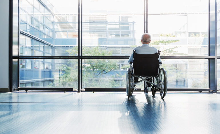 Senior Man in Wheelchair looking out of a window in a care facility corridor